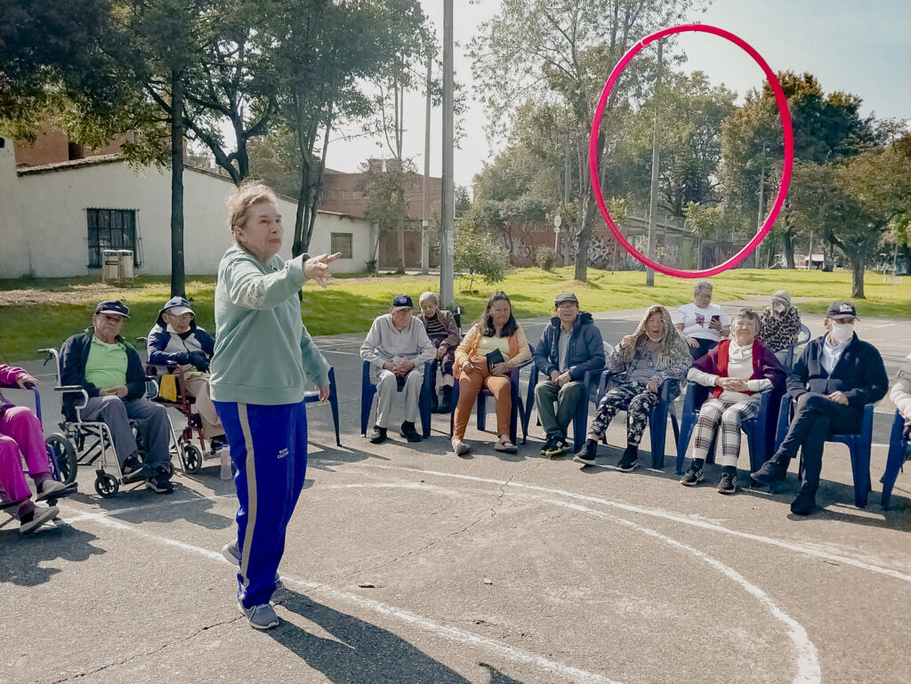 Adultos mayores participando en una actividad deportiva al aire libre en Bogotá, usando herramientas como aros, balones y bandas elásticas en un parque soleado.